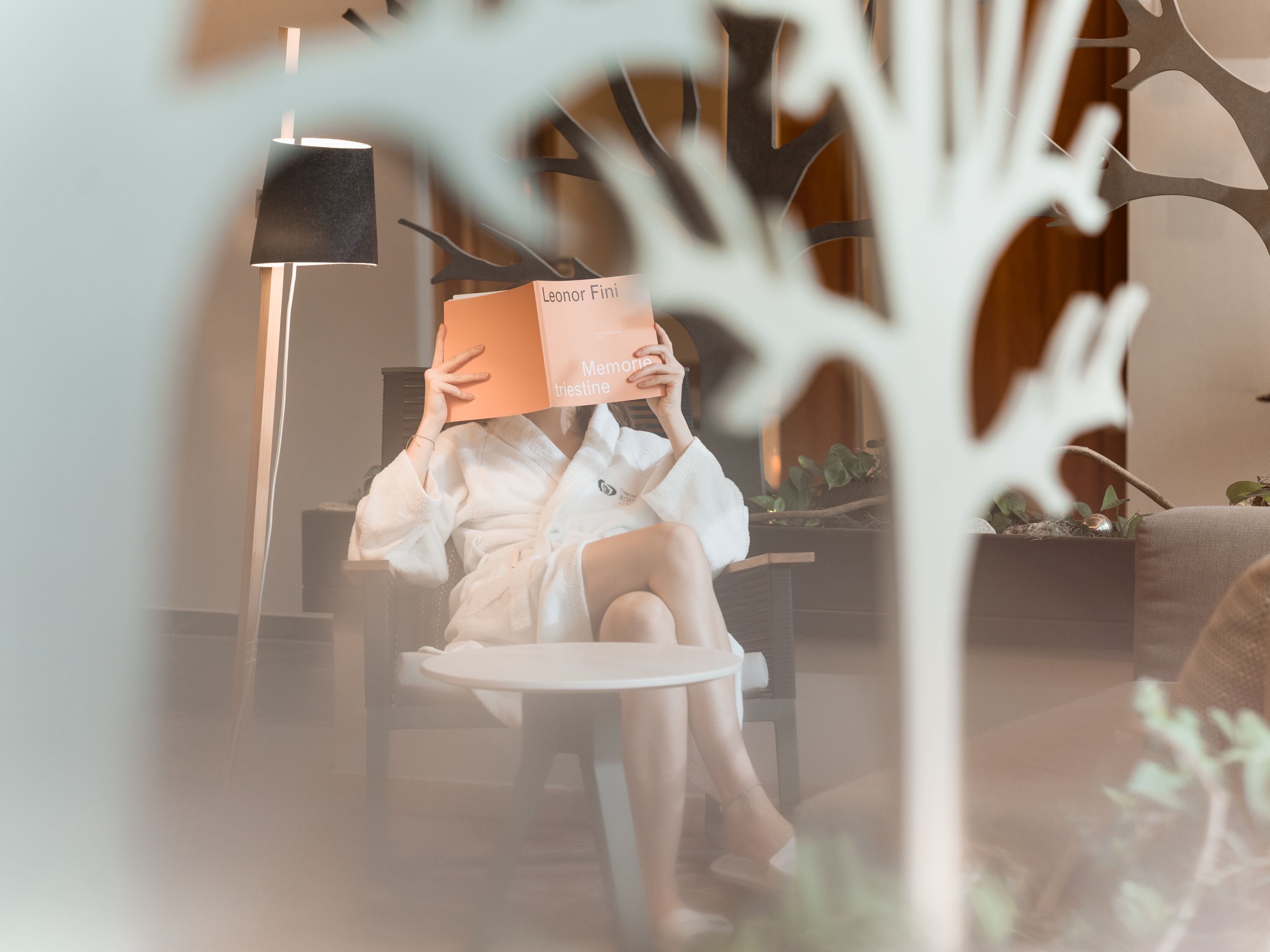 A person sitting on a chair in a room with a round table, a lamp, and a plant.