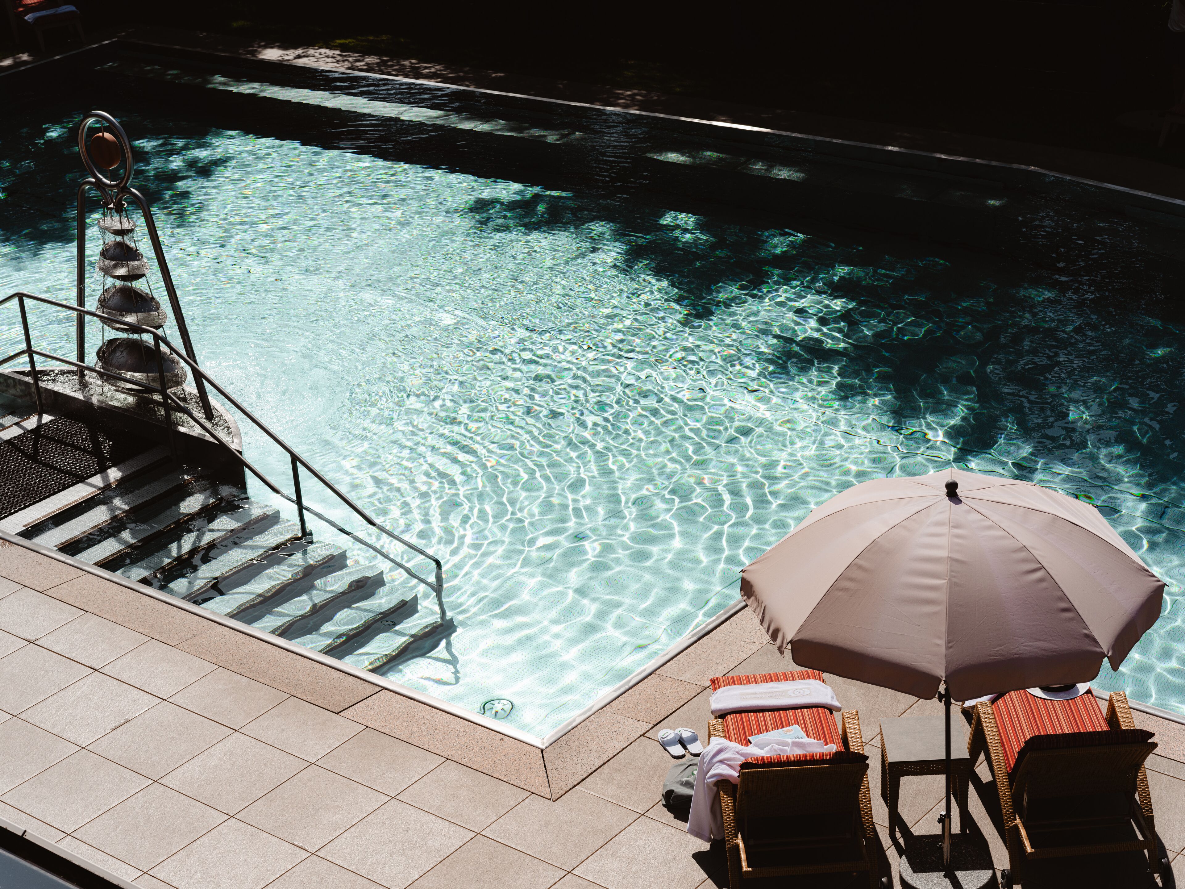 A poolside area with clear water, a set of stairs leading into the pool, and two lounge chairs with an umbrella.