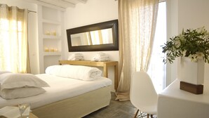 A hotel room with a bed, a bedside table with a lamp, a mirror, a chair, and a window with curtains. - Kapetan Tasos Suites (Milos)