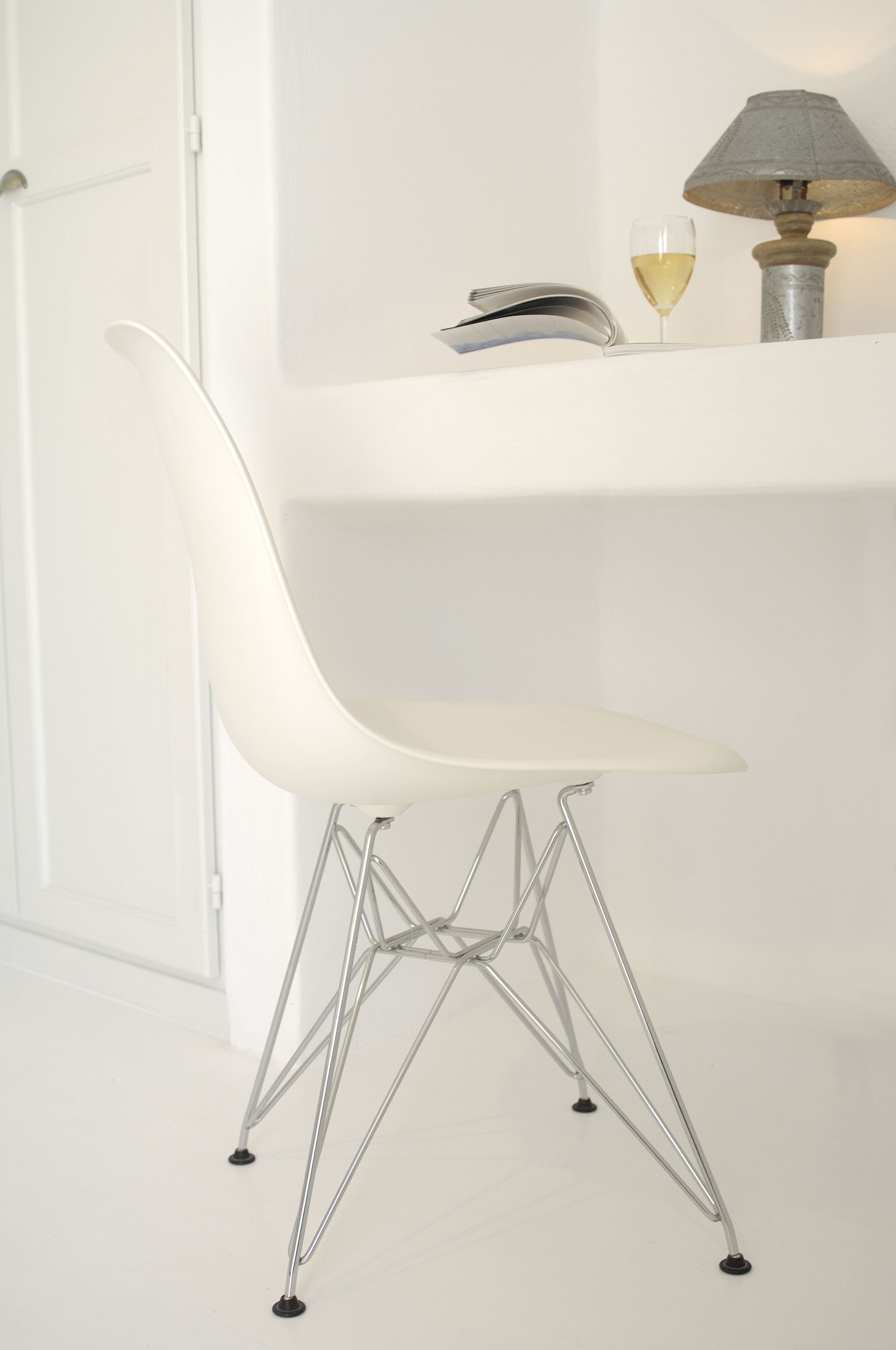 Une chaise blanche avec une structure en métal, un verre de vin blanc et une lampe posée sur une surface blanche.