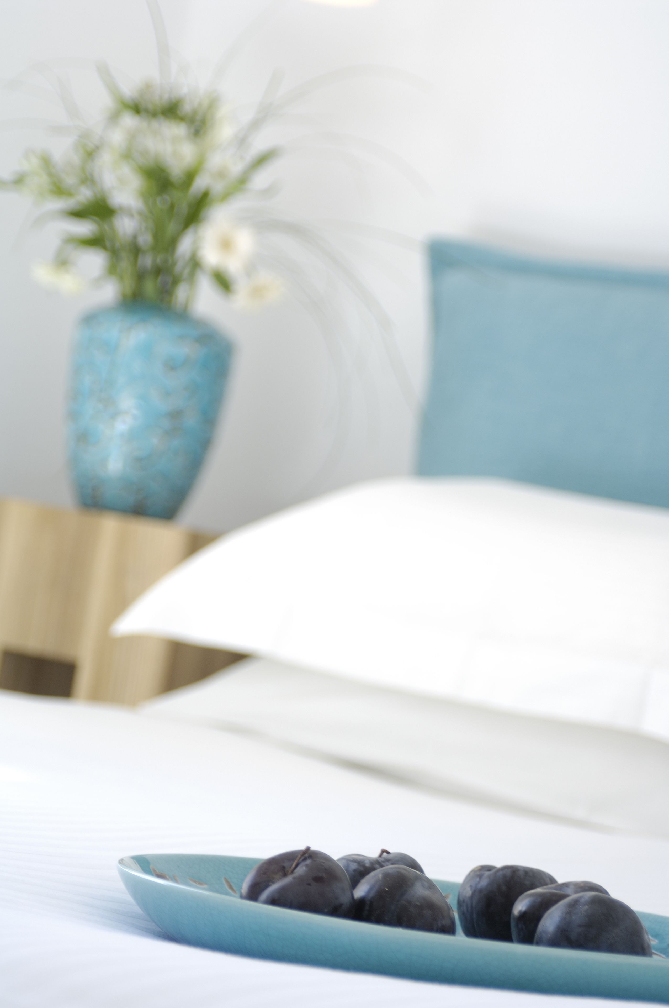 A bed with white bedding, a blue pillow, and a blue vase with flowers on a bedside table.
