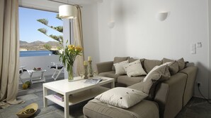 A modern living room with a beige sofa, a white coffee table, and a view of the sea through large windows. - Kapetan Tasos Suites (Milos)