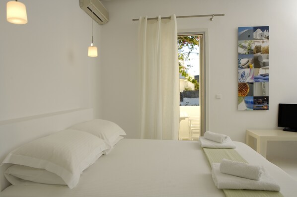 A hotel room with a bed, white curtains, a painting on the wall, and a TV on a side table. - Kapetan Tasos Suites (Milos)