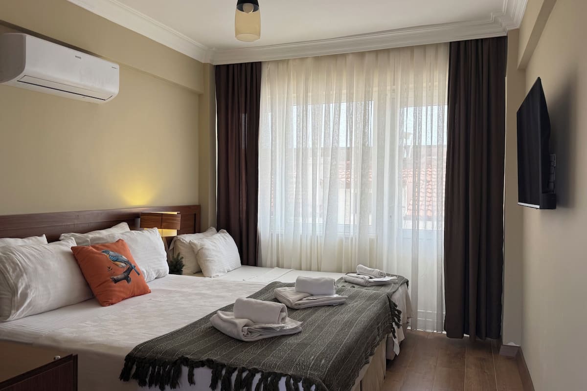 A hotel room with a bed, a flat-screen TV, an air conditioning unit, a window with curtains, and a bedside table with a lamp.