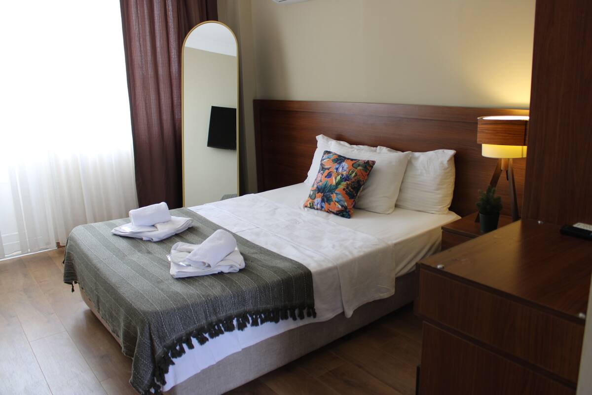 A hotel room with a bed, a bedside table, a lamp, a mirror, and a window with curtains.