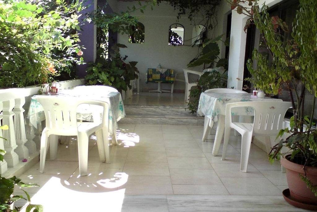 An outdoor patio area with white plastic chairs and tables, surrounded by potted plants and a tiled floor.