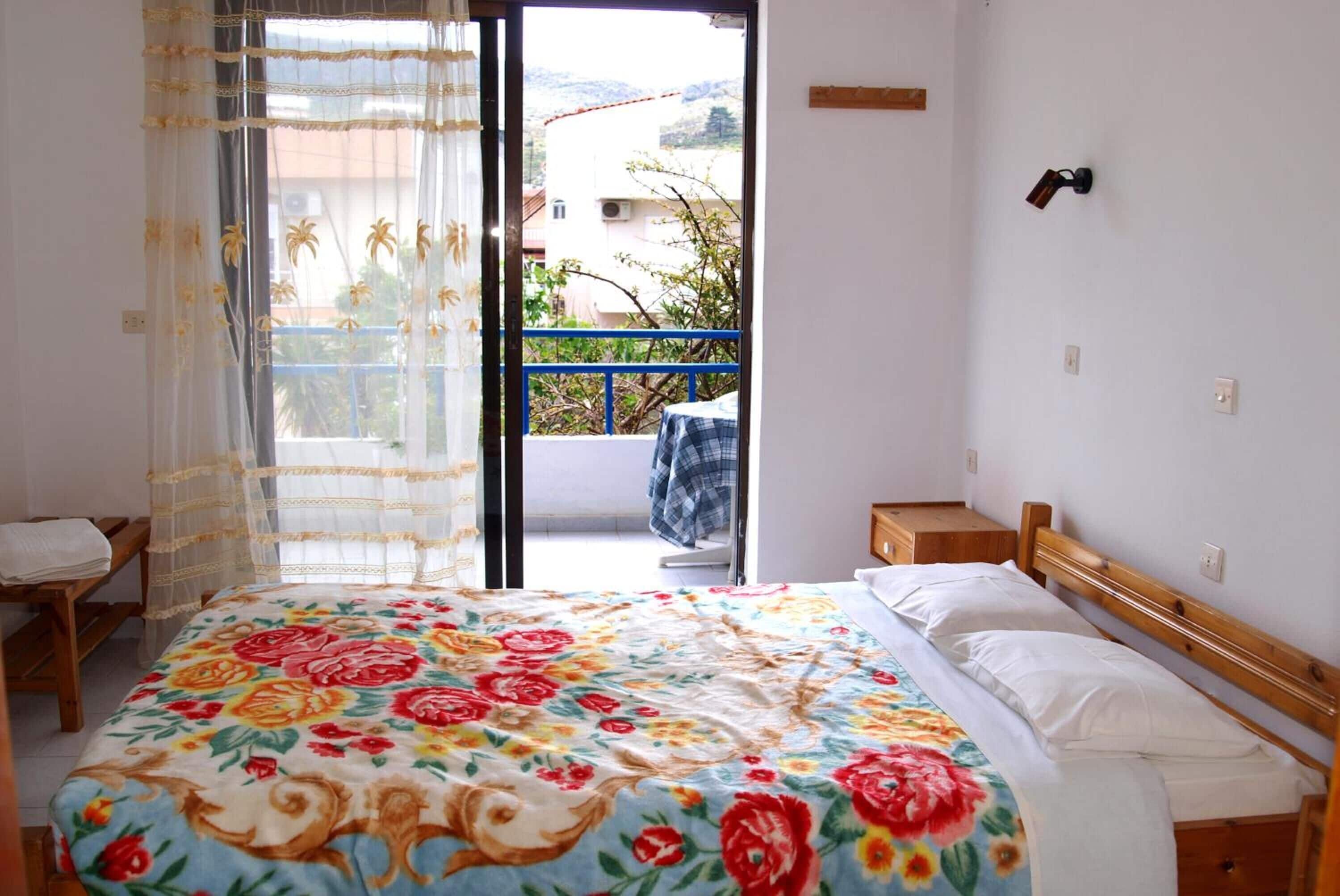 Un dormitorio con cama, una silla, un balcón con barandilla azul y vistas a edificios y árboles.