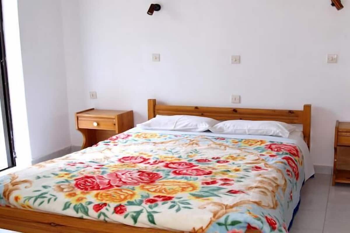 A bedroom with a wooden bed, white pillows, a floral bedspread, and a nightstand with a vase of flowers.
