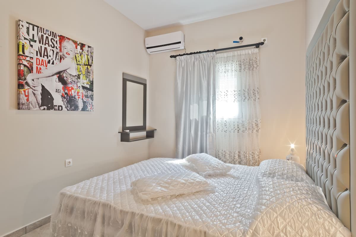 A bedroom with a bed, a wall-mounted air conditioner, a decorative mirror, and a window with sheer curtains.