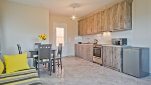 A compact kitchen with wooden cabinets, a refrigerator, and a microwave. There is a dining area with a table and chairs. - Montemar Studios & Apartments (Karpathos)