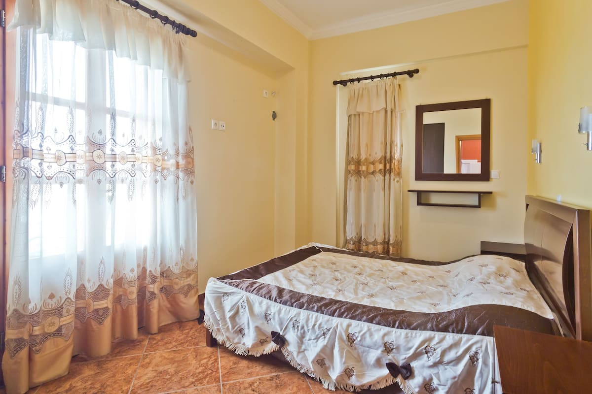 A bedroom with a bed, curtains, a mirror, and a tiled floor.
