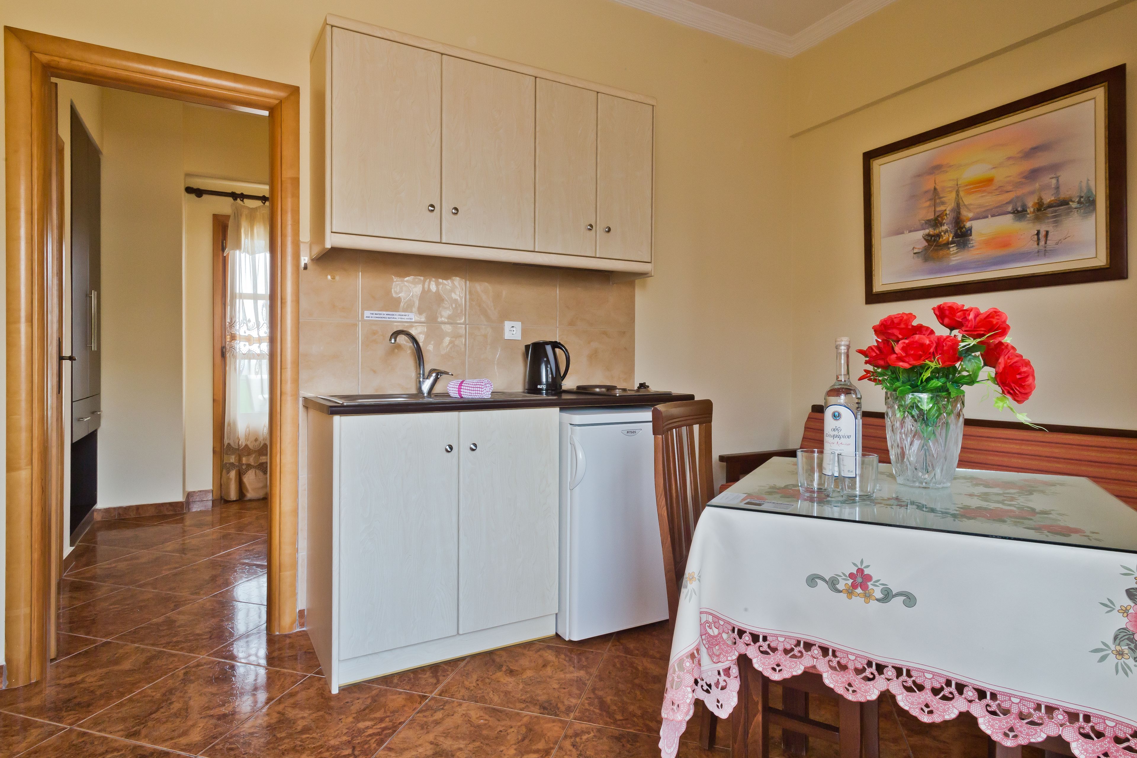 A compact kitchen with white cabinets, a small refrigerator, and a dining table with a floral tablecloth.