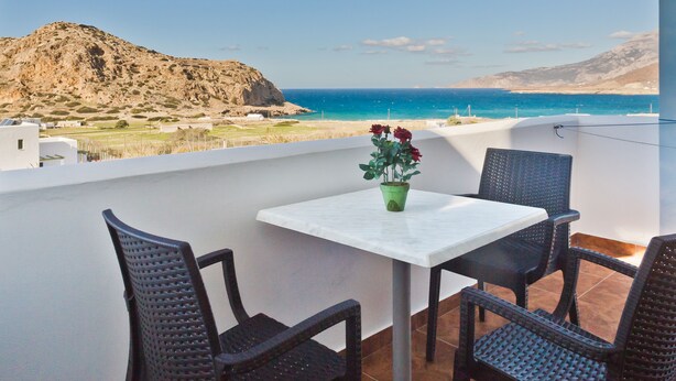A balcony with a table and chairs, overlooking a coastal landscape with a mountain and the sea. - Montemar Studios & Apartments (Karpathos)