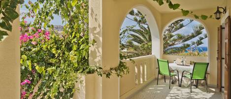 A covered outdoor patio with a table and chairs, surrounded by arches and greenery.