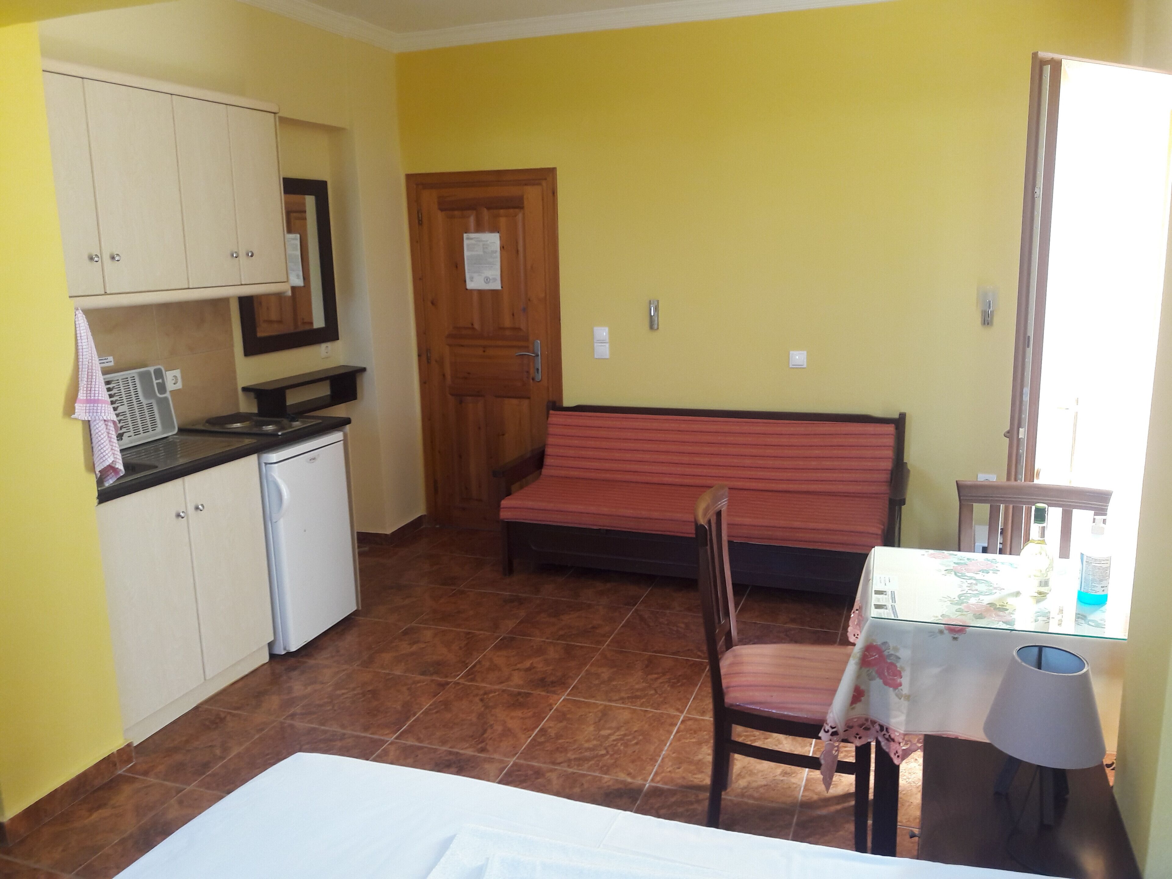 A compact room with a kitchenette, a wooden door, a bench, a table with a lamp, and a bed.