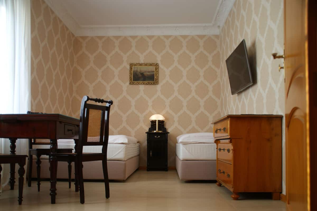 Una habitación de hotel con un escritorio de madera, dos camas, una mesita de noche con lámpara, un aparador y un televisor en la pared.
