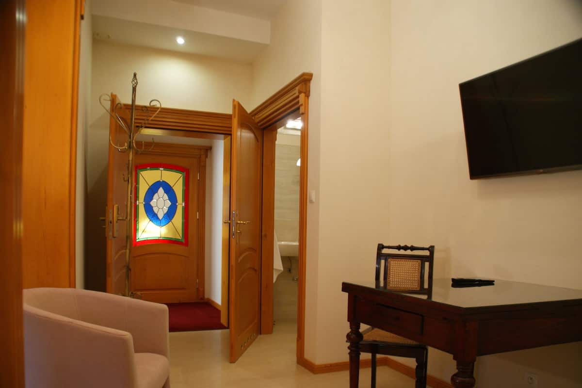 Una habitación con un escritorio de madera, una silla, un televisor montado en la pared y una puerta que da al baño.