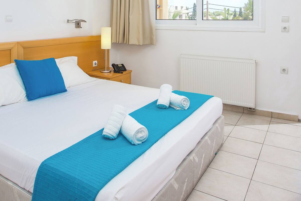 Una cama bien tendida con ropa de cama blanca y azul, una manta de color azul y toallas enrolladas. Hay dos mesitas de noche con lámparas y un teléfono. La habitación tiene un cabecero de madera, una ventana con cortinas y un radiador.