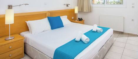 A neatly made bed with white and blue bedding, a blue throw blanket, and rolled towels. There are two bedside tables with lamps and a phone. The room has a wooden headboard, a window with curtains, and a radiator.
