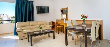 A modern living room with a sofa, coffee table, dining table, and chairs. A flat-screen TV is mounted on the wall, and there is a window with curtains.