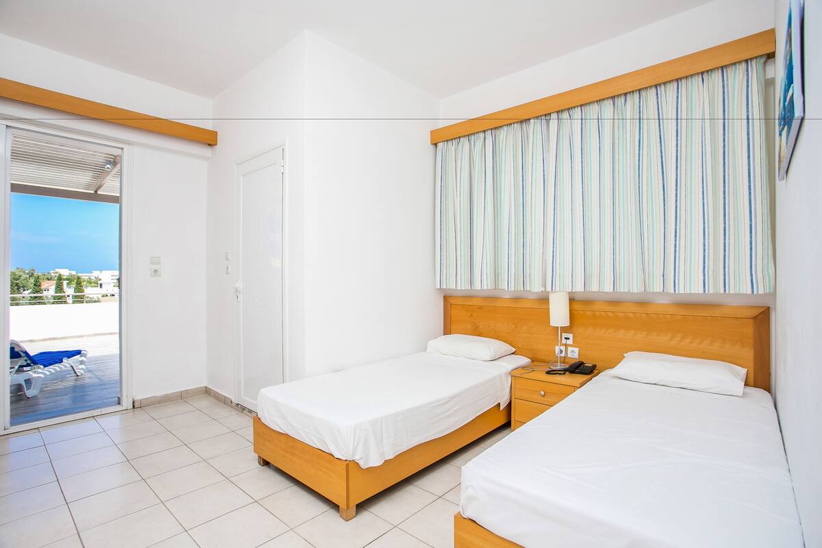Habitación de hotel con dos camas, cabecero de madera, cortina a rayas, mesita de noche con lámpara y puerta corrediza de cristal que da a un balcón.