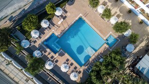 Une vue aérienne d’une piscine rectangulaire entourée de chaises longues et de parasols, avec des arbres et un chemin pavé à proximité.