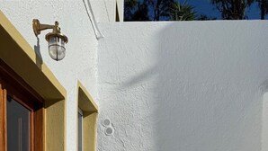 Un mur extérieur blanc avec une porte en bois, un banc blanc avec des coussins et une table blanche sur laquelle se trouve un seul objet.