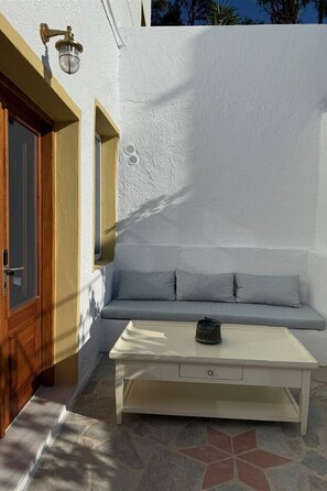 A white exterior wall with a wooden door, a white bench with cushions, and a white table with a single object on it.