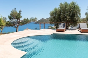 Piscina con sillones de descanso y una pérgola, con vista a un cuerpo de agua y colinas.