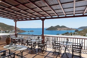 A terrace with tables and chairs, overlooking a coastal landscape with hills and the sea.