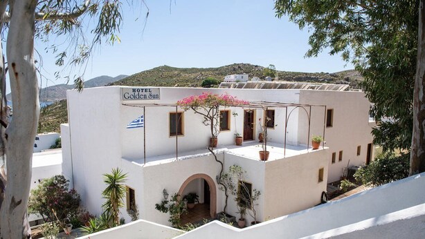 Un bâtiment blanc sur lequel on peut lire l’inscription « HÔTEL Golden Sun ». Il possède un balcon orné de plantes en pot et offre une vue sur une colline.