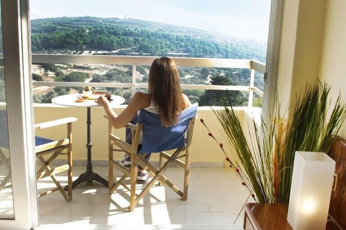 Un balcon avec une table et des chaises, une personne assise, et une vue sur un paysage verdoyant.