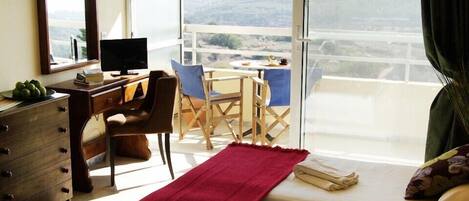 Habitación de hotel con escritorio, una silla, televisor, cama con dos almohadas, una manta roja y balcón con mesa y sillas.