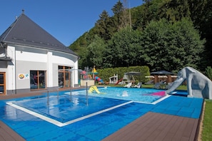 Una piscina con tobogán, rodeada de sillones de descanso y un edificio con tejado inclinado.