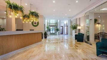 A modern hotel lobby with a reception desk, a large green wreath, and a glass elevator.