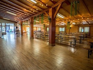A spacious hall with wooden floors and tables, featuring exposed wooden beams and hanging plants.