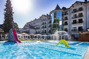 Una piscina con toboganes y una fuente, rodeada de sillones de descanso y sombrillas.