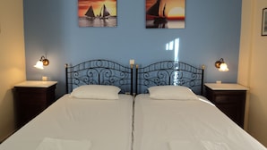 Un lit double avec une tête de lit en métal noir, deux tables de chevet avec des lampes et deux tableaux représentant des voiliers au coucher du soleil, accrochés au mur.