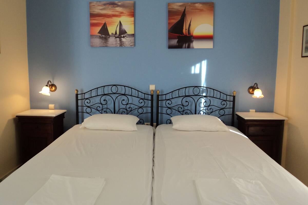 Una cama doble con cabecera de metal negro, dos mesitas de noche con lámparas, y dos cuadros en la pared que muestran veleros al atardecer.