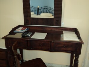 Un bureau en bois, avec un miroir au-dessus, qui reflète un lit doté d’une tête de lit bleue. On peut voir des papiers et une télécommande sur le bureau.