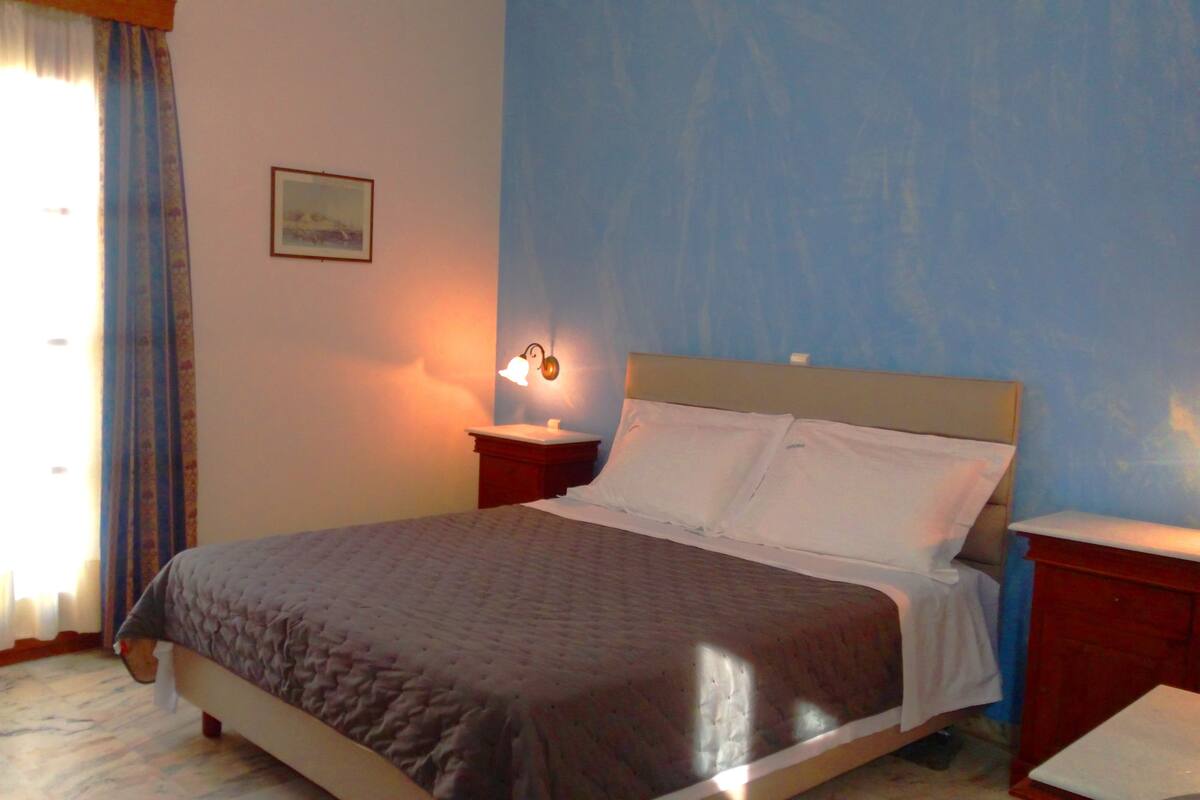 Un dormitorio con cama, mesita de noche, lámpara de pared y ventana con cortinas.