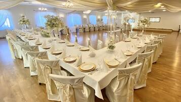 Un salón de banquetes con largas mesas dispuestas para un evento formal, con manteles blancos, platos con detalles dorados y sillas blancas decoradas con lazos.