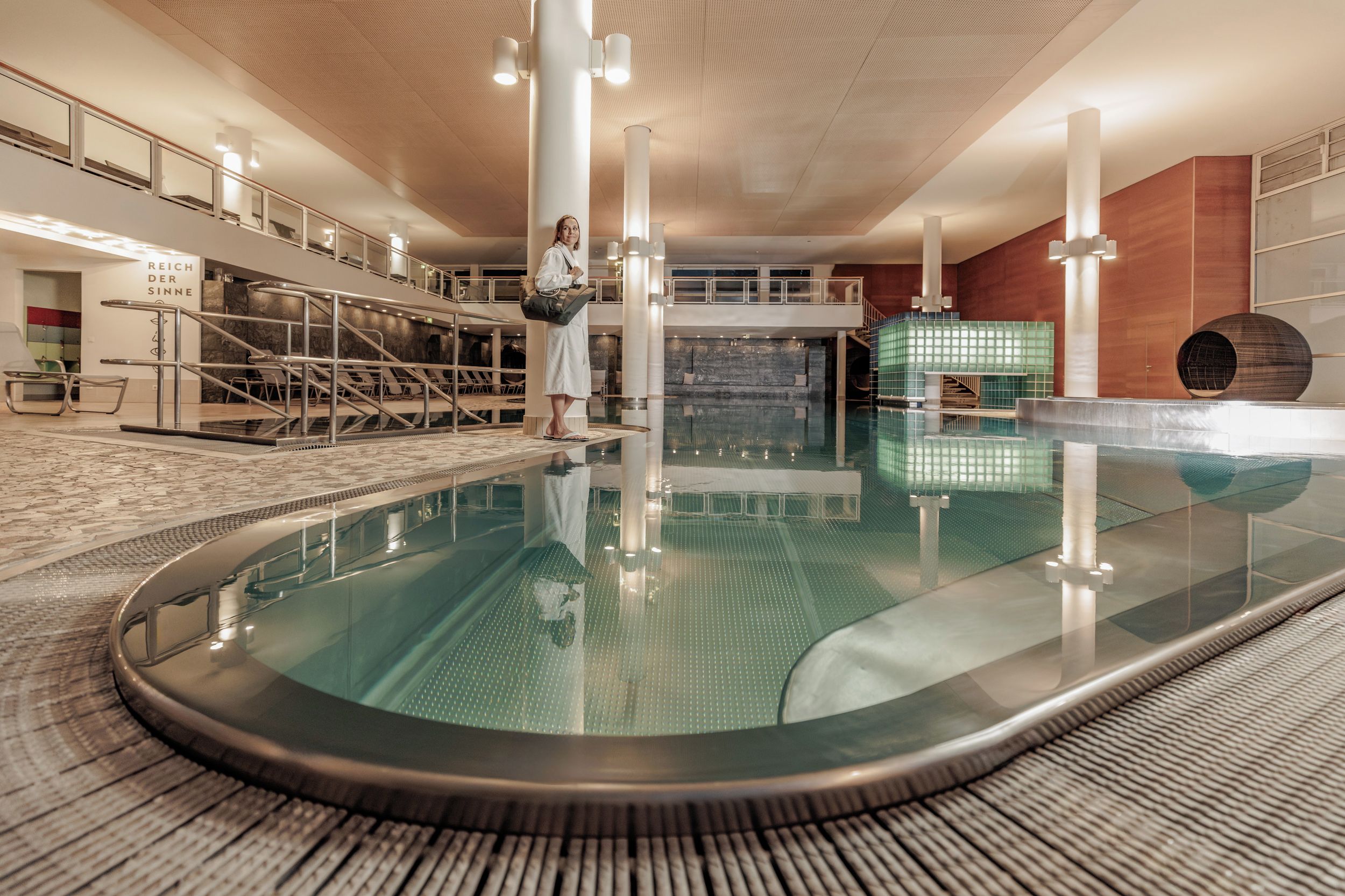A modern indoor swimming pool with a glass barrier, a lounge area with seating, and a staircase leading to an upper level.