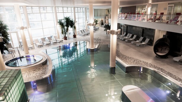 A spacious indoor pool area with lounge chairs and a hot tub, surrounded by large windows and indoor plants. - Reduce Hotel Vital (Bad Tatzmannsdorf)