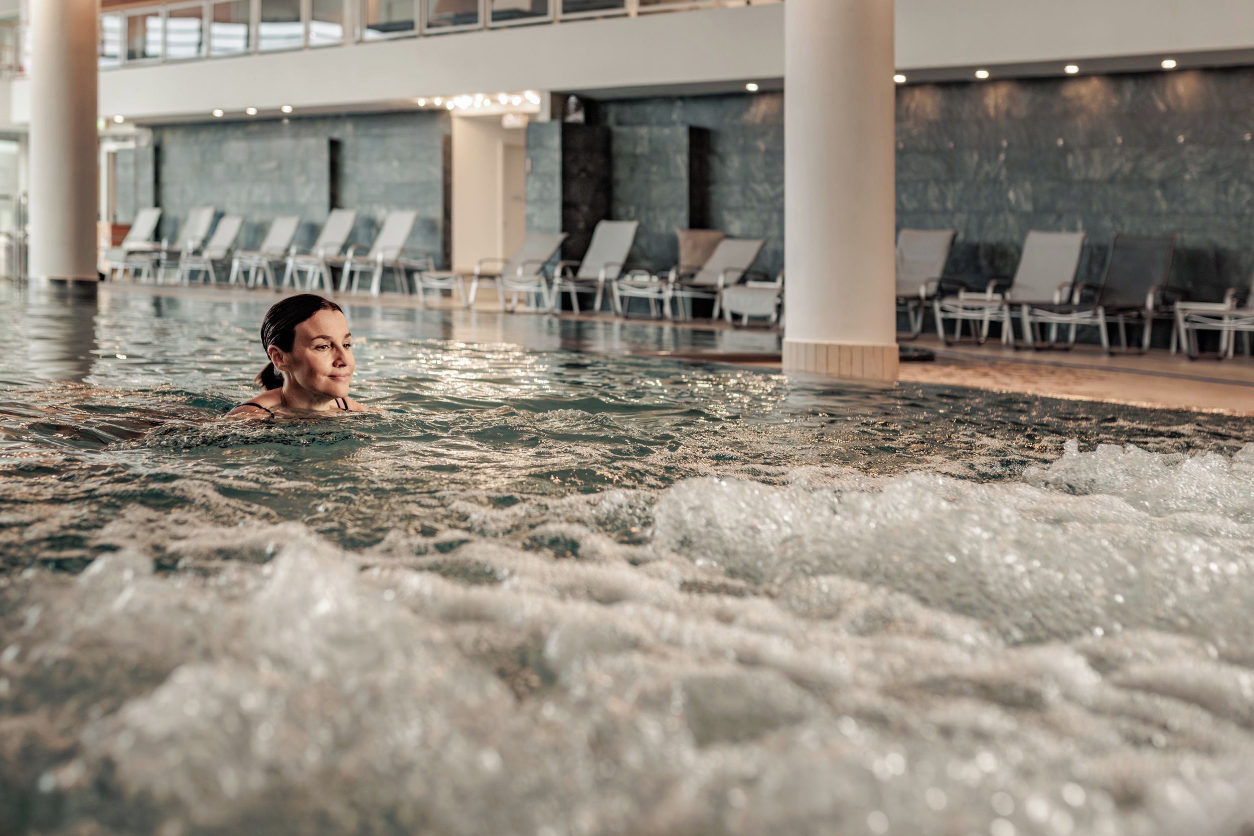 A person in a hot tub with water jets, surrounded by lounge chairs and a tiled wall.