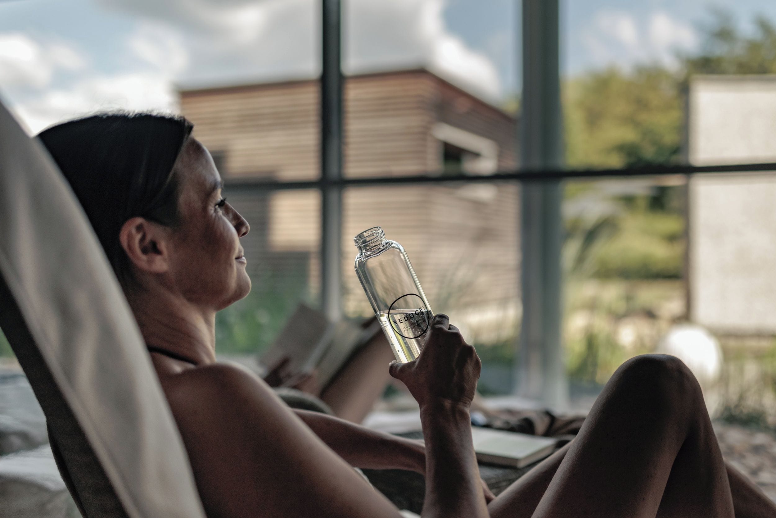 Una persona recostada en una habitación iluminada por el sol, sosteniendo una botella transparente.