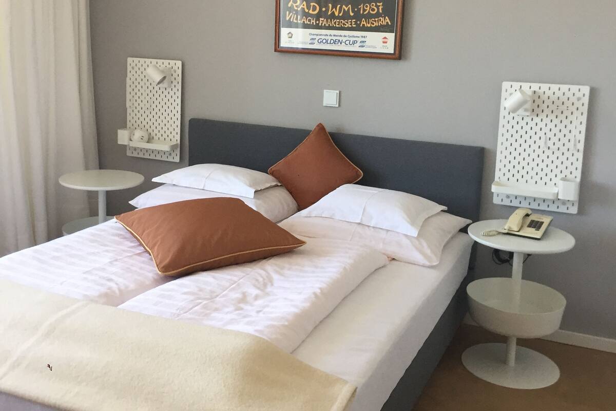 A neatly made bed with white and brown pillows, a small bedside table with a lamp, and a framed picture on the wall.