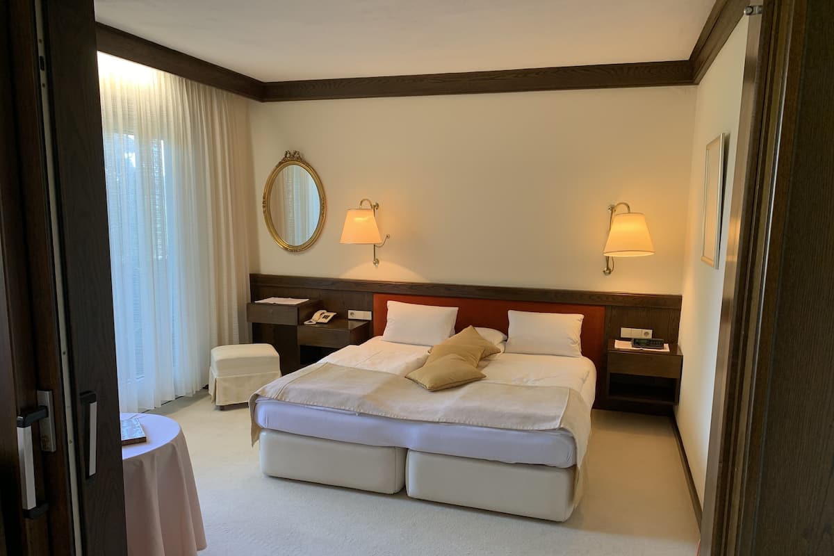 A hotel room with a large bed, two bedside tables, a nightstand, a round table, and a window with curtains.