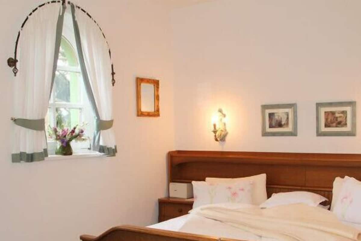 A bedroom with a wooden bed, a bedside table, a window with curtains, and wall-mounted lights.