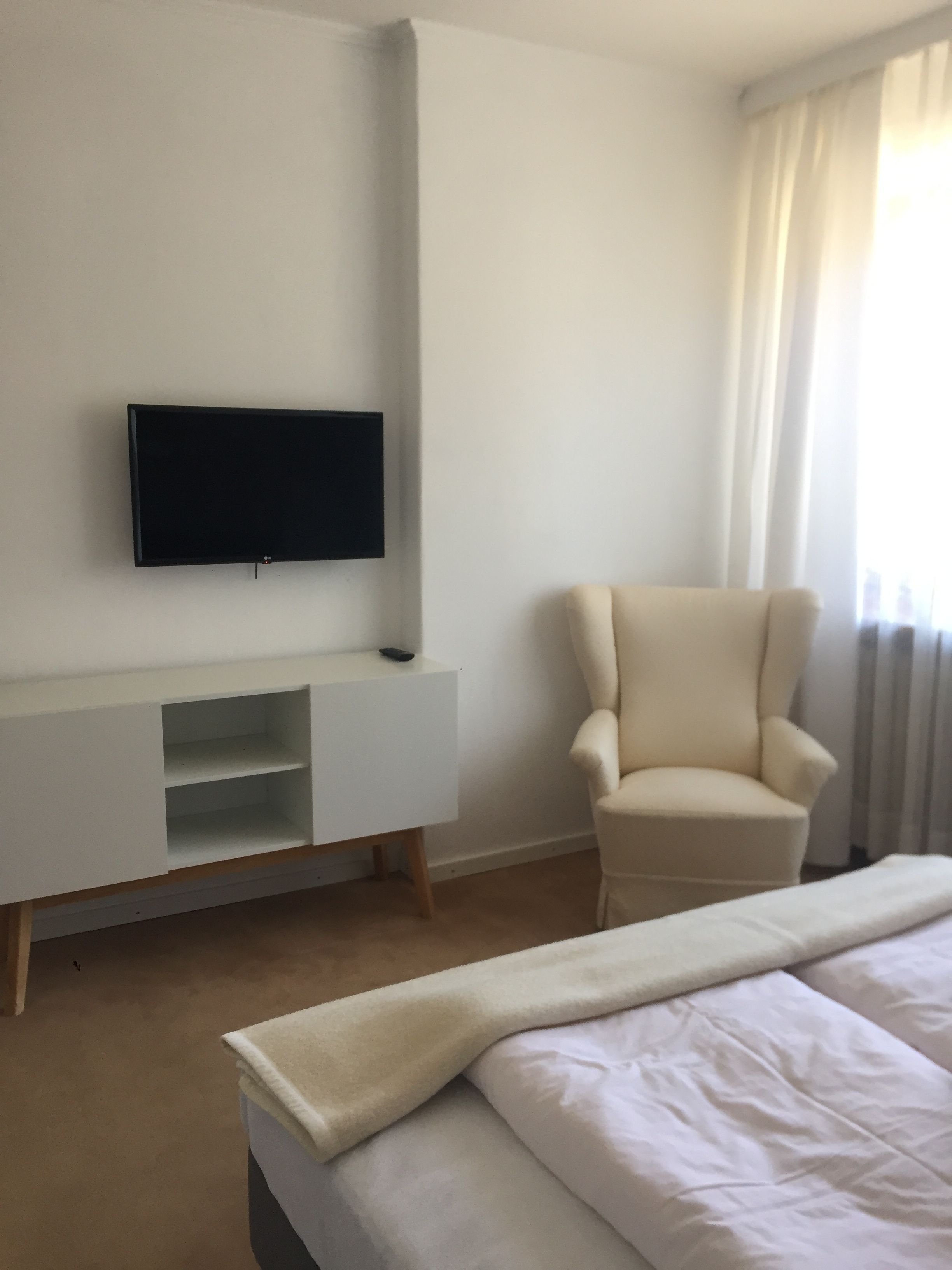 A hotel room with a bed, a TV mounted on the wall, a chair, and a window with curtains.